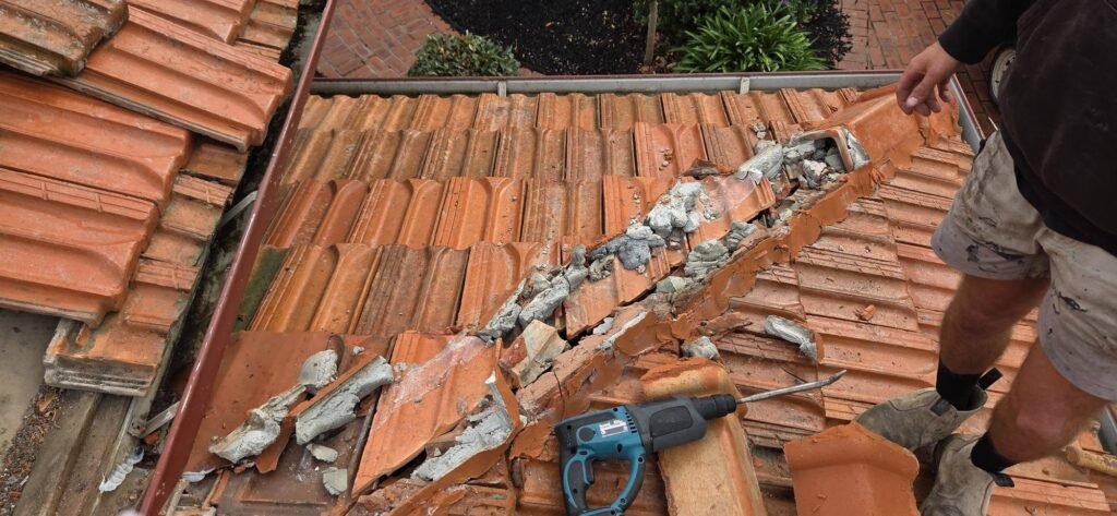 Roofer chipping out old ridge capping and mortar on a terracotta tiled roof during a roof leak repair in Essendon